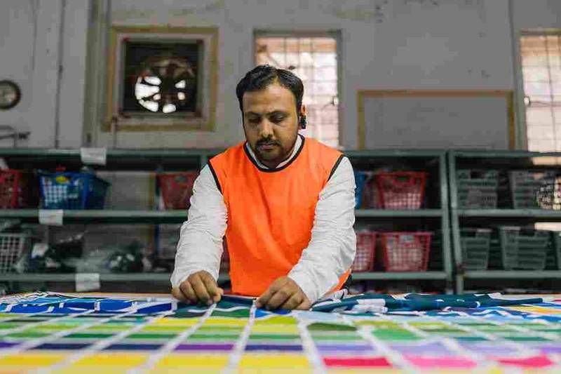 A man in an orange safety vest carefully examines colorful fabric samples on a table in a textile manufacturing facility.