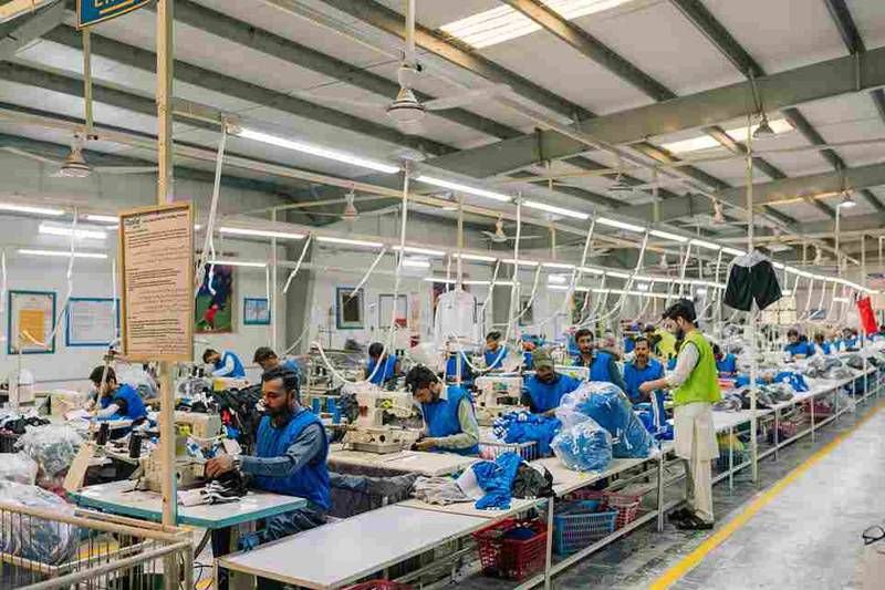 A busy textile manufacturing facility with several workers dressed in blue uniforms operating sewing machines and organizing fabric. The environment is well-lit and equipped for garment production.